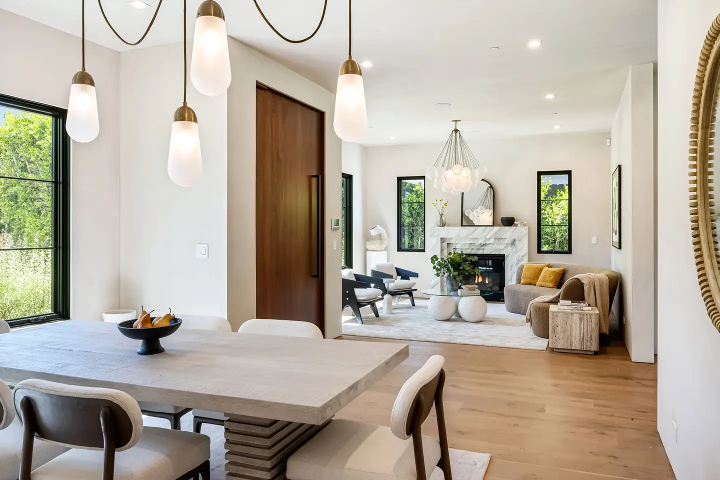 Staged open plan dining and living room with a light oak table, sculptural pendants and a marble fireplace
